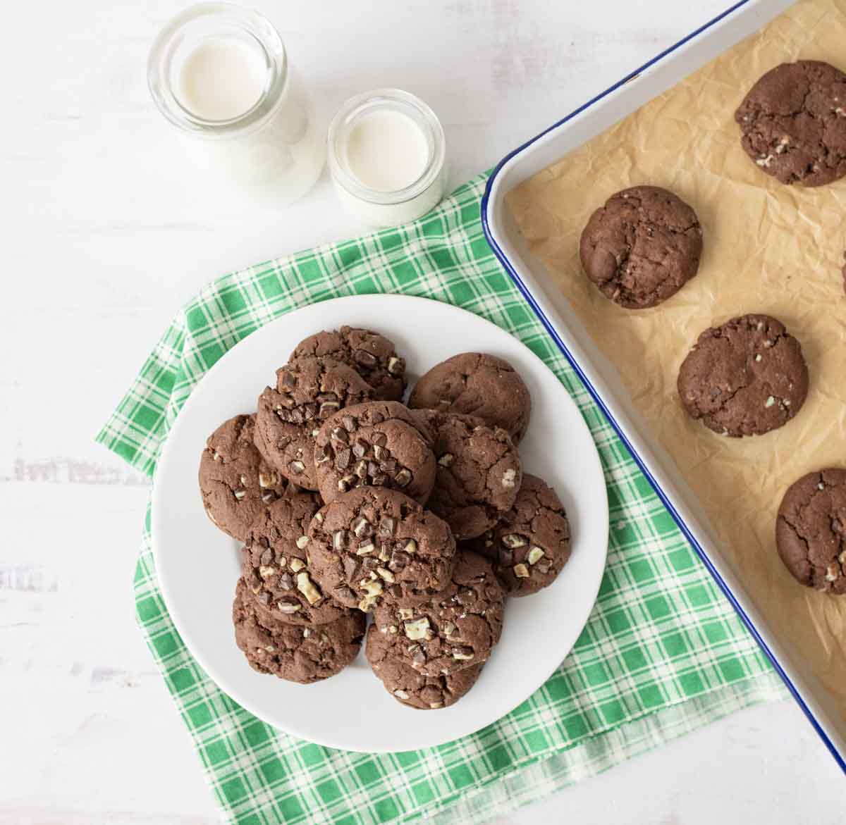 A plate of chocolate chip cookies on a green plaid cloth, next to a baking tray with more cookies and two small bottles of milk on a white surface.