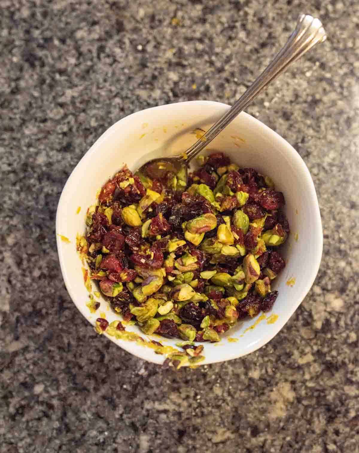 A white bowl filled with a colorful mixture of chopped pistachios, dried cranberries, and orange zest sits on a speckled gray countertop, with a silver spoon resting inside.