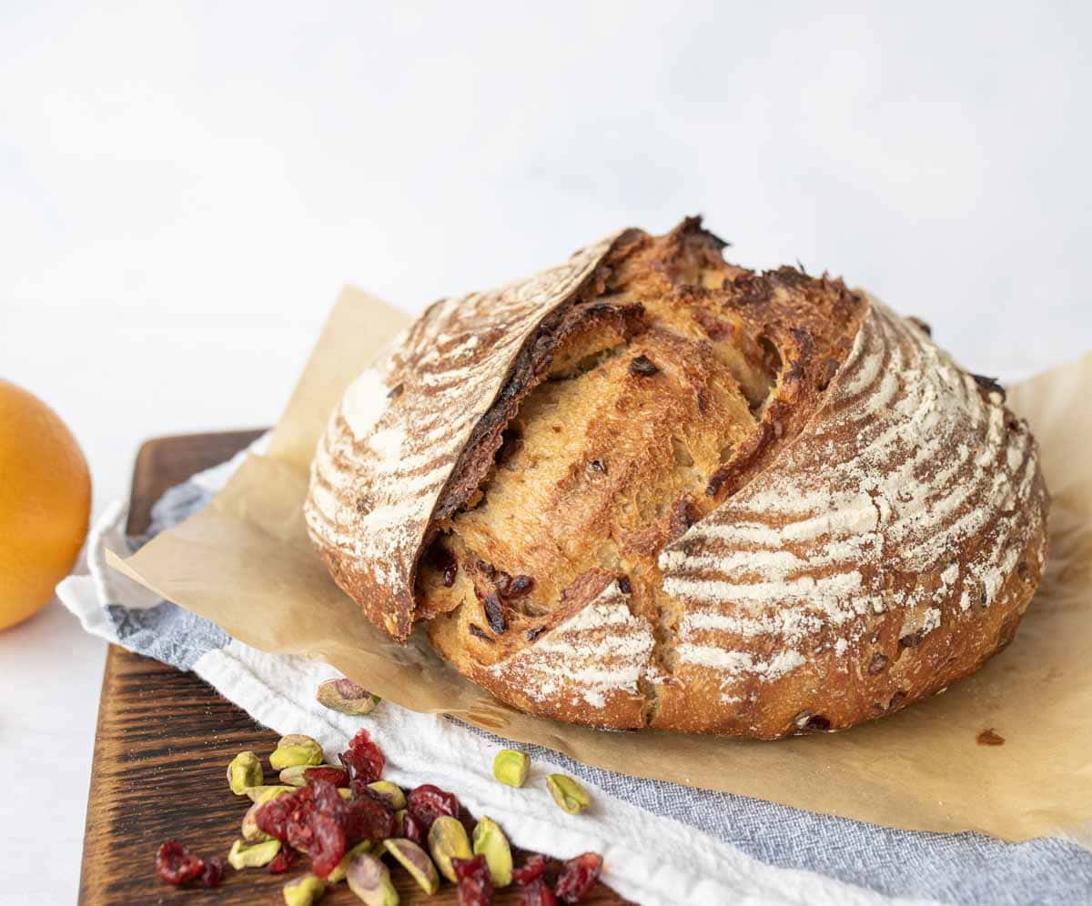 A round, rustic loaf of sourdough bread with a cracked, golden crust sits on parchment paper atop a wooden board. Nearby are scattered dried cranberries, pistachios, and part of an orange.