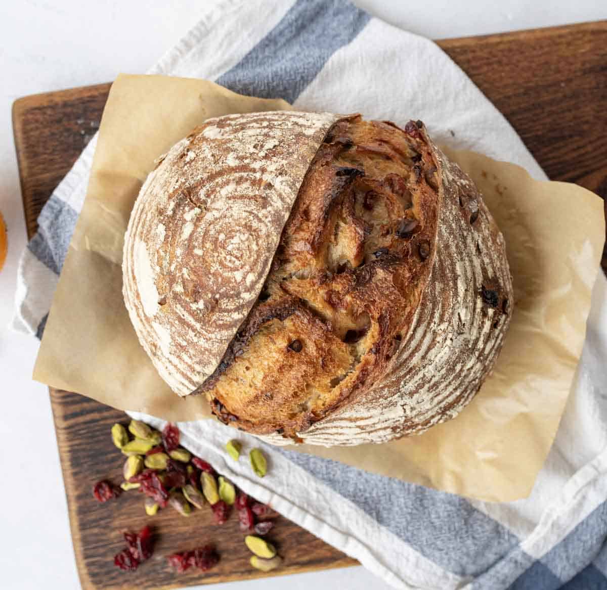 A round loaf of artisan bread sits on parchment paper atop a wooden board, with a blue and white towel underneath. Dried cranberries and green pistachios are scattered nearby.