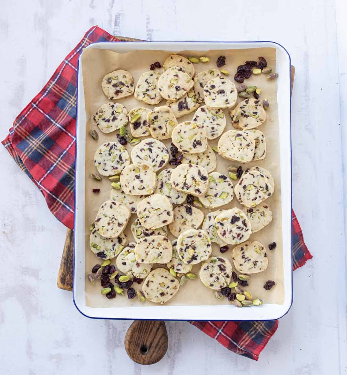 A baking tray lined with parchment paper holds sliced cookies sprinkled with pistachios and dried cranberries, resting on a red plaid cloth atop a light-colored surface.