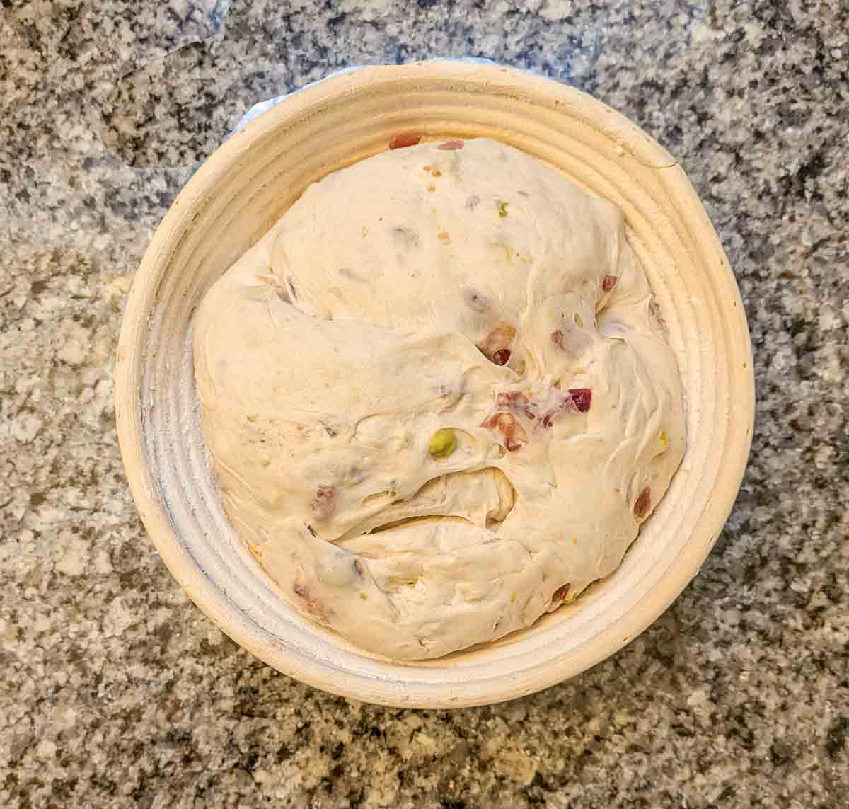 A round bowl filled with risen bread dough containing visible pieces of nuts and dried fruit, placed on a granite countertop.
