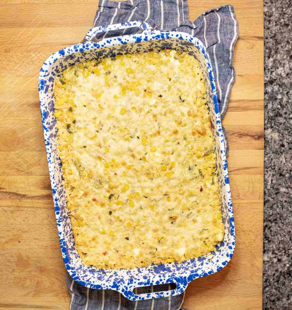 A baked corn casserole in a blue and white speckled rectangular dish sits on a wooden surface with a striped kitchen towel underneath. The casserole has a golden, cheesy top.