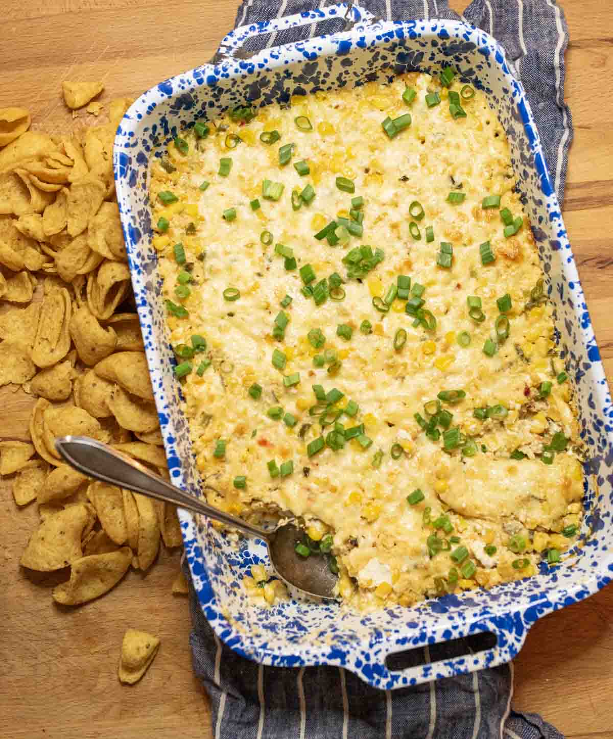 A blue and white speckled baking dish filled with creamy corn dip, topped with chopped green onions. A spoon rests in the dish, and corn chips are scattered on the wooden surface alongside.