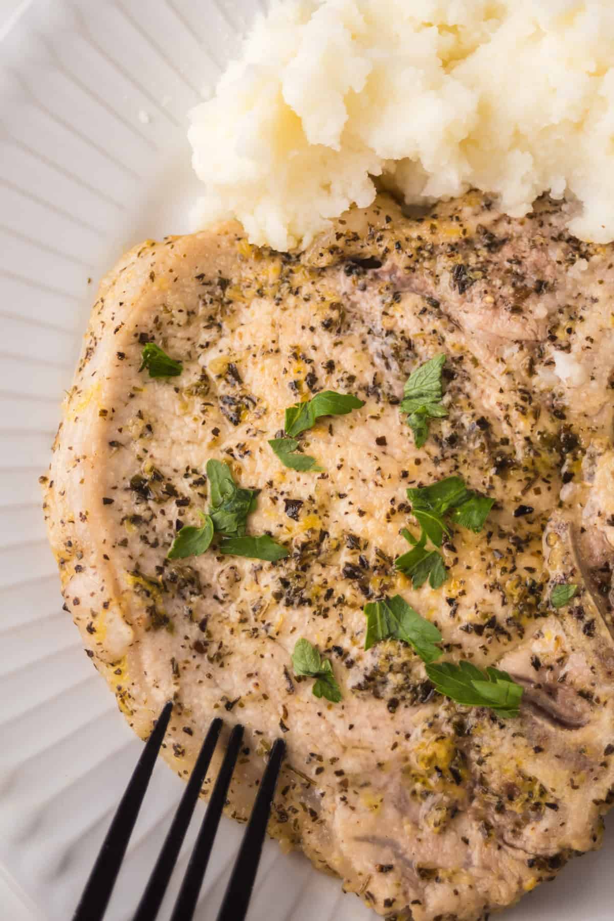 A cooked lemon pepper pork chop seasoned with herbs, garnished with fresh parsley, served on a white plate with mashed potatoes. A black fork is placed near the pork chop.