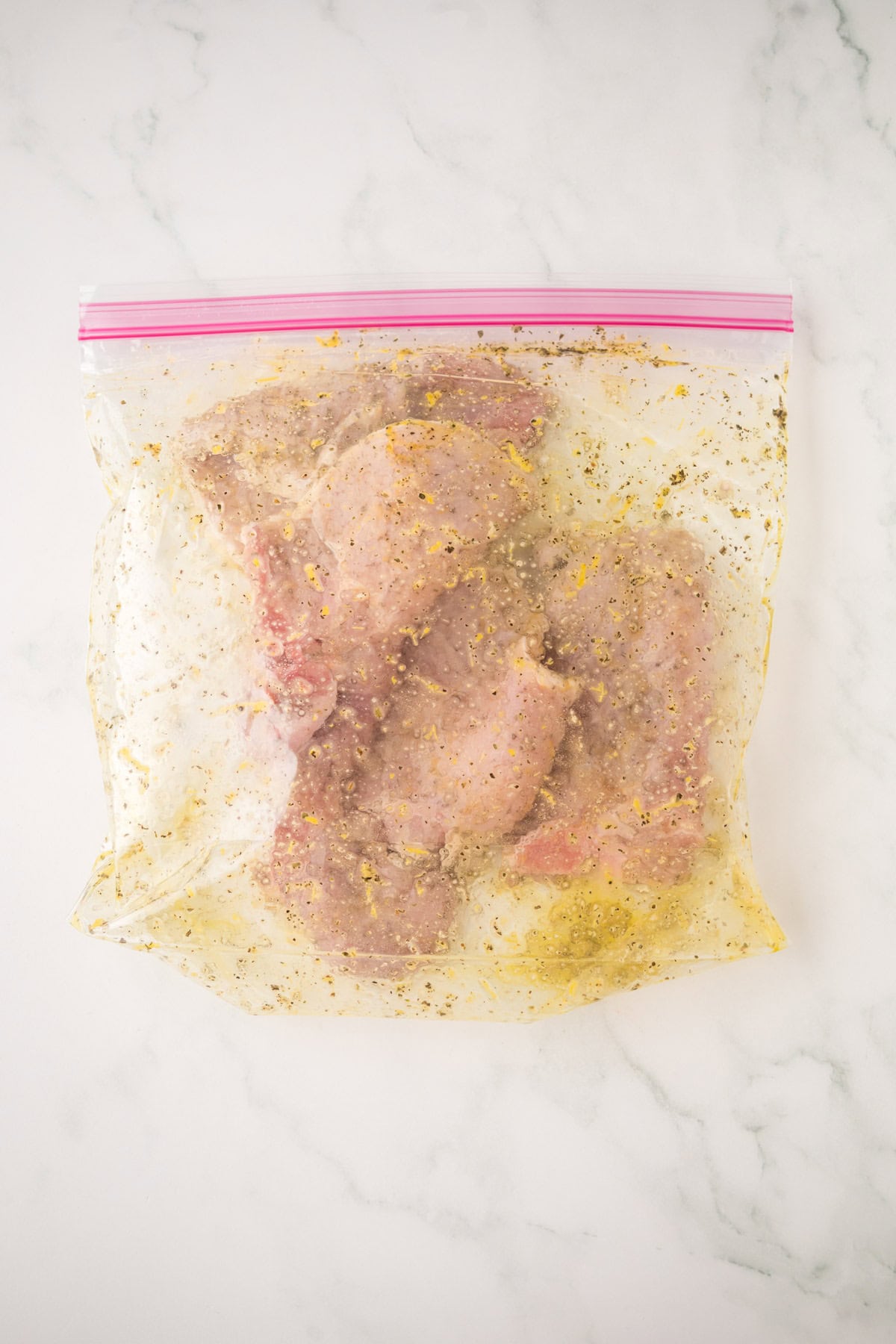 Raw chicken pieces marinating with herbs and seasoning in a clear plastic zip-top bag, reminiscent of lemon pepper pork chops, placed on a white marble surface.