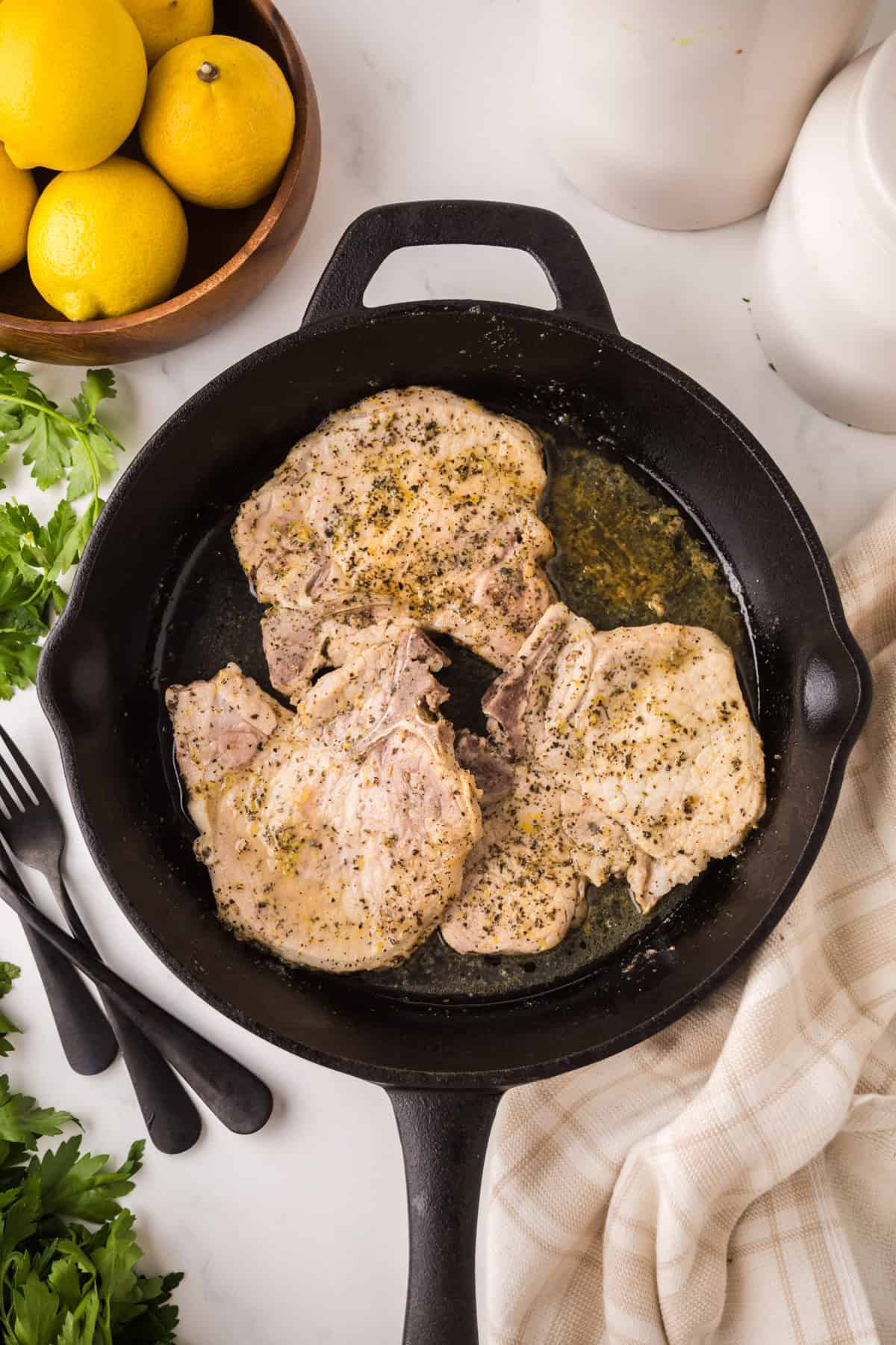Three seasoned lemon pepper pork chops sizzle in a cast iron skillet, surrounded by fresh parsley, a bowl of lemons, utensils, and a beige kitchen towel on a white countertop.