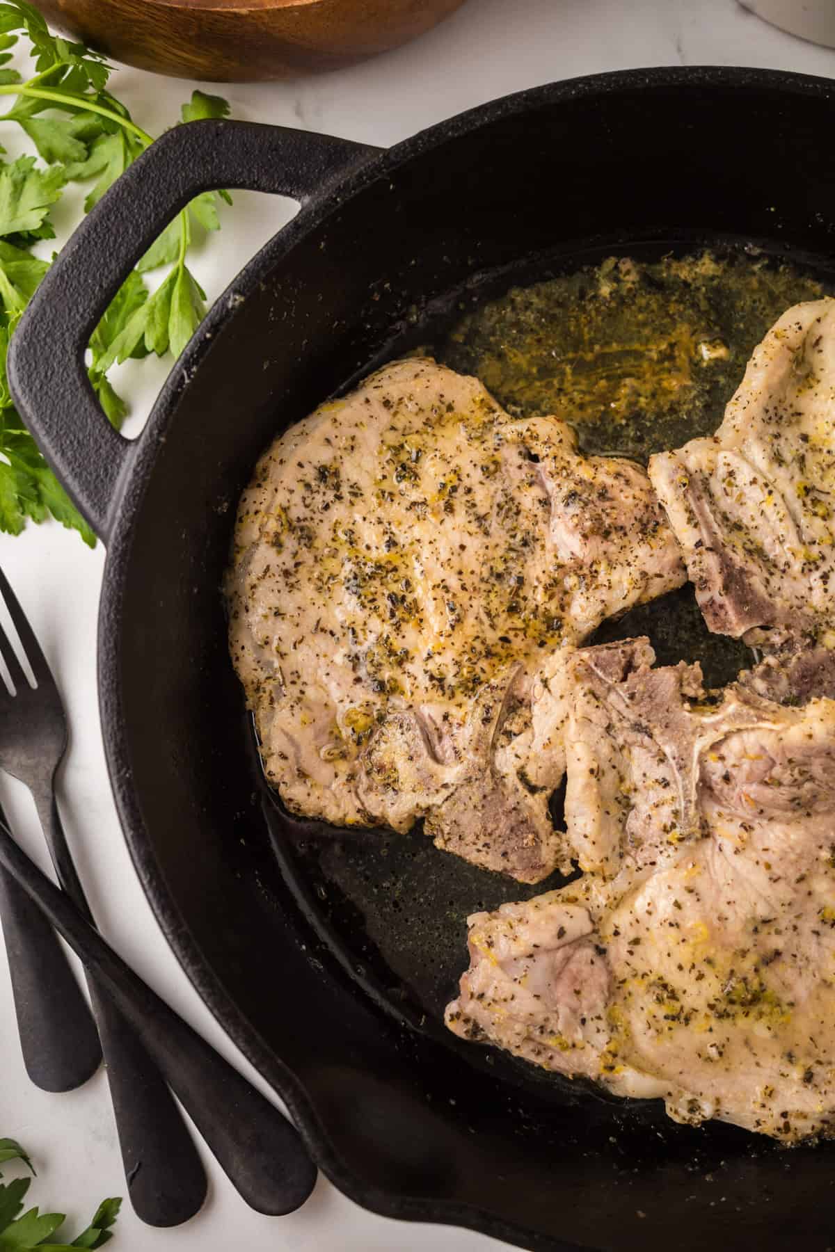 Three lemon pepper pork chops sizzle in a black cast iron skillet, herbs sprinkled on top. Fresh parsley and cutlery rest on the white surface beside the pan.