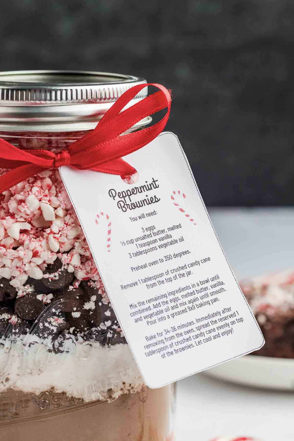 A glass jar filled with brownie mix layers, topped with crushed peppermint and chocolate chips, is tied with a red ribbon and a recipe tag labeled Peppermint Brownies. A brownie is blurred in the background.