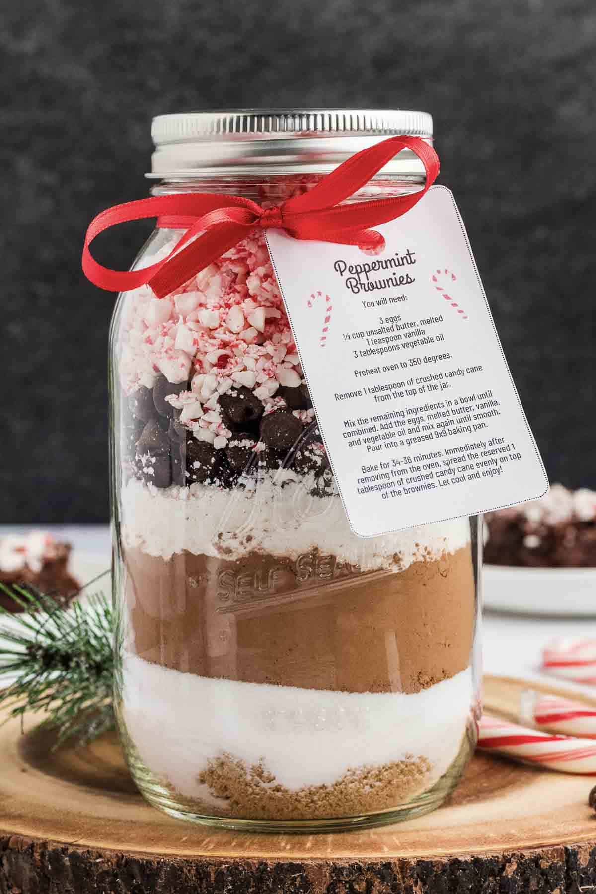 A mason jar filled with layered brownie mix ingredients—brown sugar, white sugar, cocoa, chocolate chips, and crushed peppermint. The jar is topped with a red ribbon and a recipe card labeled Peppermint Brownies.