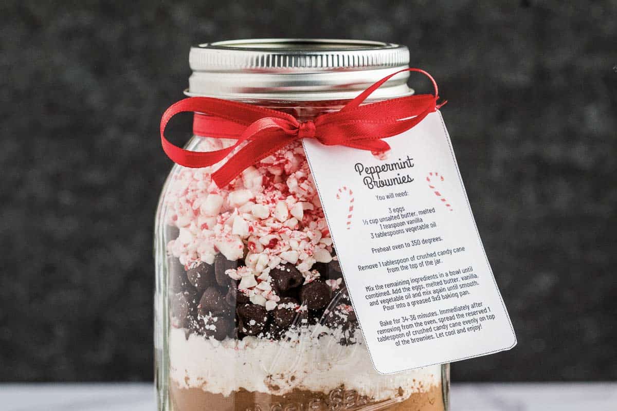 A mason jar filled with layered brownie mix ingredients, topped with crushed peppermint, chocolate chips, and flour, tied with a red ribbon and a recipe tag labeled Peppermint Brownie.