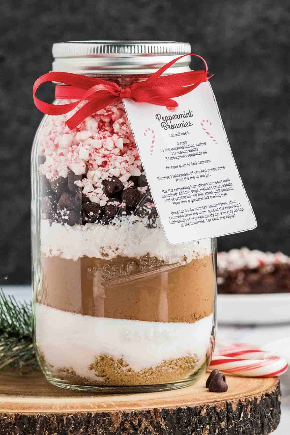 A mason jar filled with layered baking ingredients for peppermint chocolate brownies, topped with crushed peppermint, chocolate chips, and marshmallows, decorated with a red ribbon and a recipe card labeled “Peppermint Brownies.”.