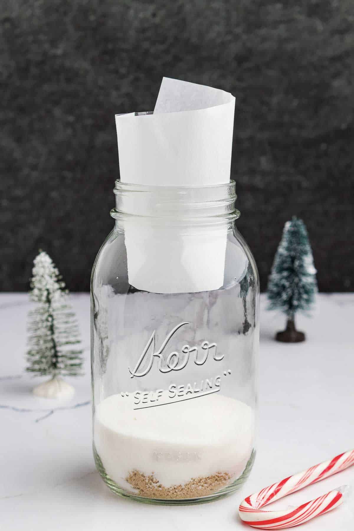 A glass Kerr mason jar containing white and brown sugar, with a rolled-up piece of parchment paper inside. The jar is on a white surface, with miniature Christmas trees and a candy cane nearby.