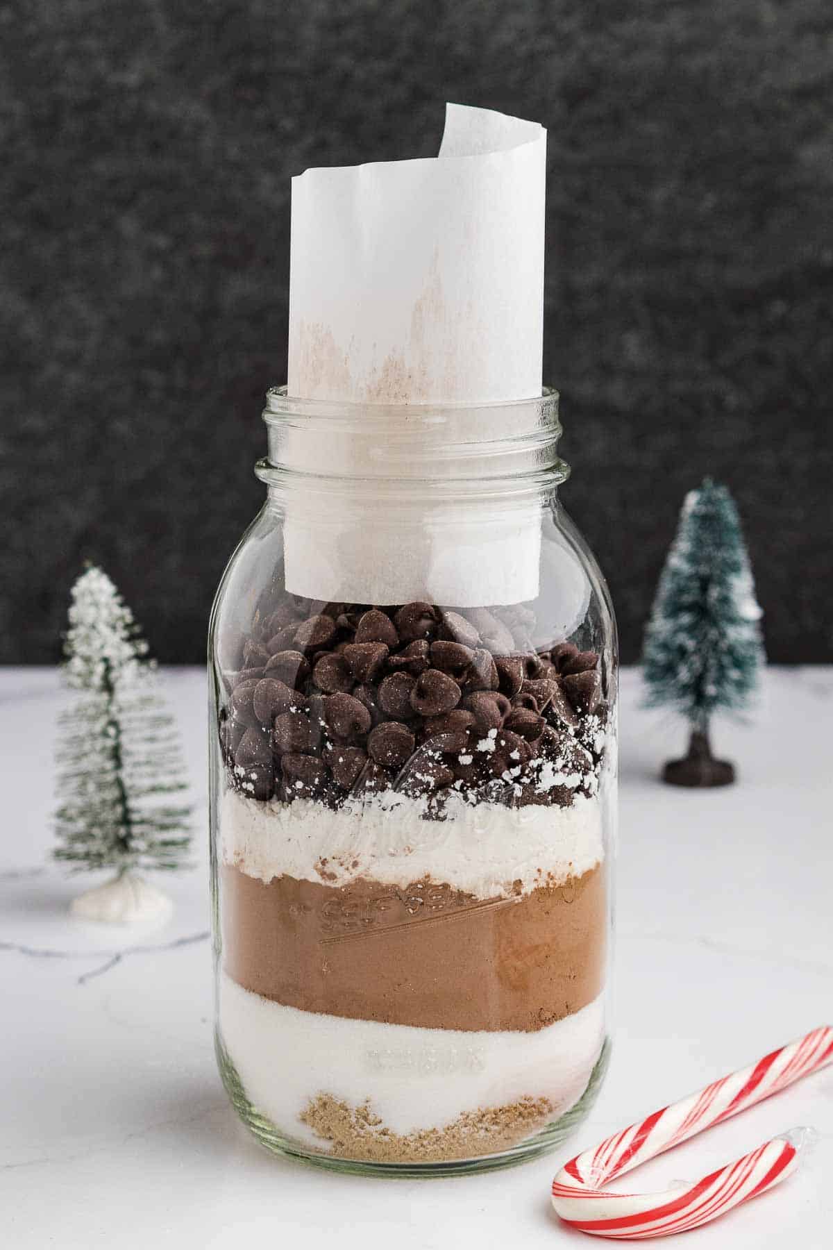 A glass jar filled with layered dry ingredients for baking, topped with chocolate chips and a rolled piece of parchment paper. Two miniature Christmas trees and a candy cane are in the background.