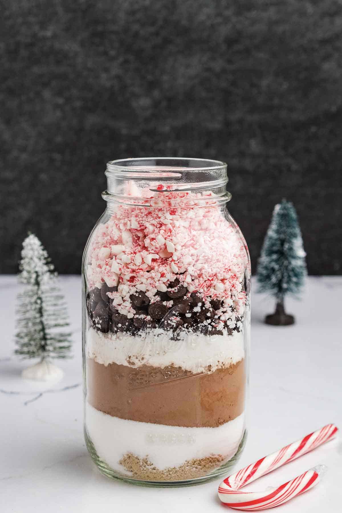 A glass jar filled with layered hot cocoa mix, including crushed peppermint, chocolate chips, cocoa powder, and sugar. Two candy canes and small decorative pine trees are in the background.