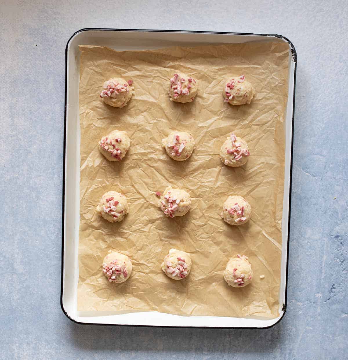 A baking tray lined with parchment paper holds twelve evenly spaced cookie dough balls topped with crushed candy pieces, ready to be baked.