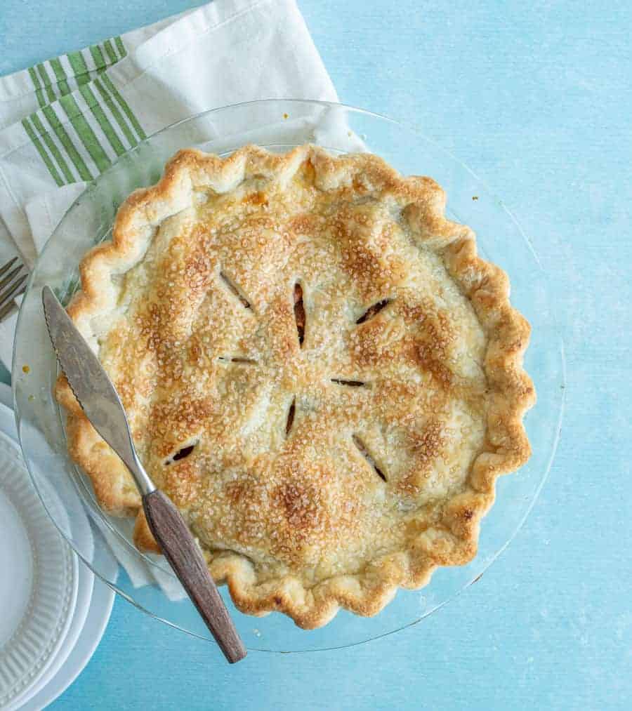 A golden-brown pie with a flaky crust—showcasing how to make pie crust—rests in a clear glass dish. A knife and server sit on top, with a white napkin featuring green stripes nearby on a light blue surface.