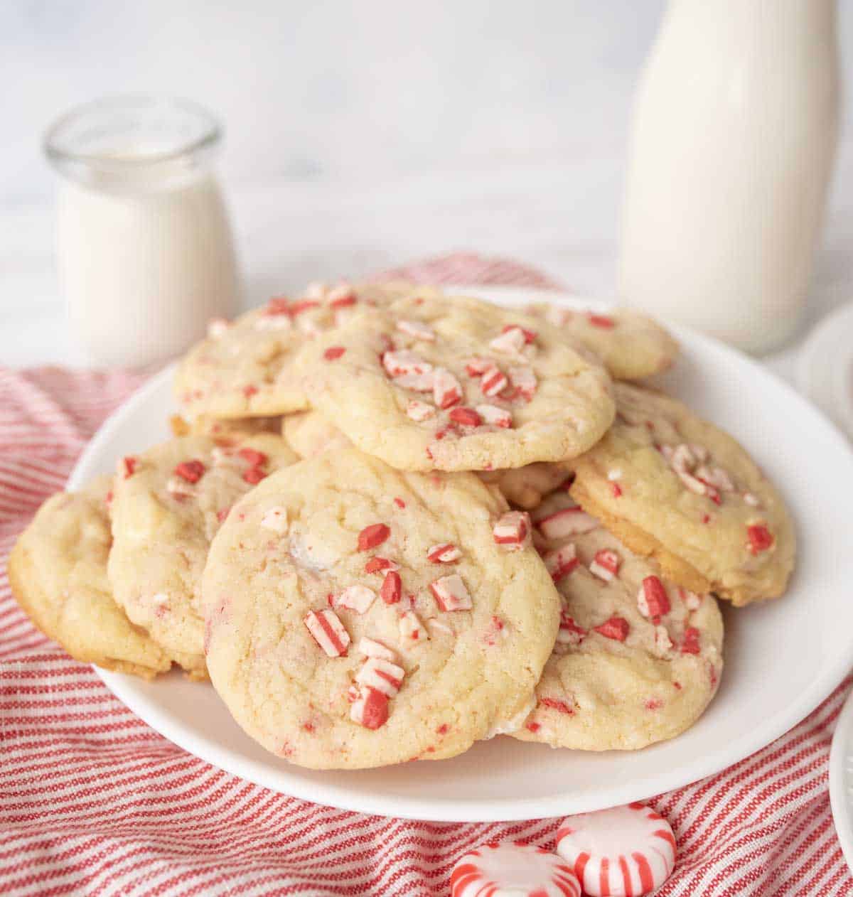 A plate of white chocolate peppermint cookies topped with crushed peppermint candies sits on a red-striped cloth, with a jar and bottle of milk in the background.
