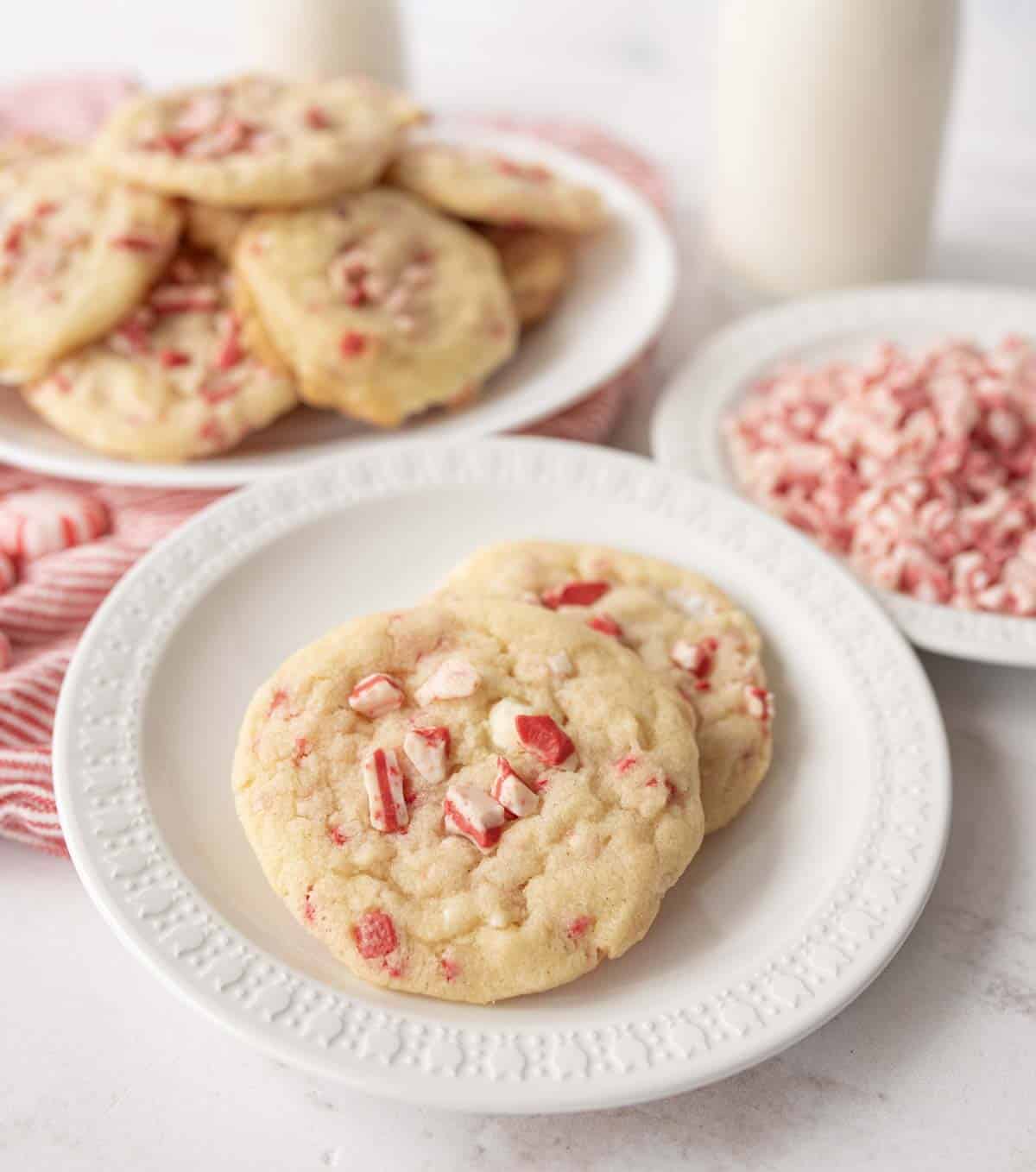 Two peppermint cookies sit on a white plate, topped with crushed peppermint candy pieces. In the background, there’s a plate of more cookies, a bowl of crushed peppermint, and two bottles of milk.