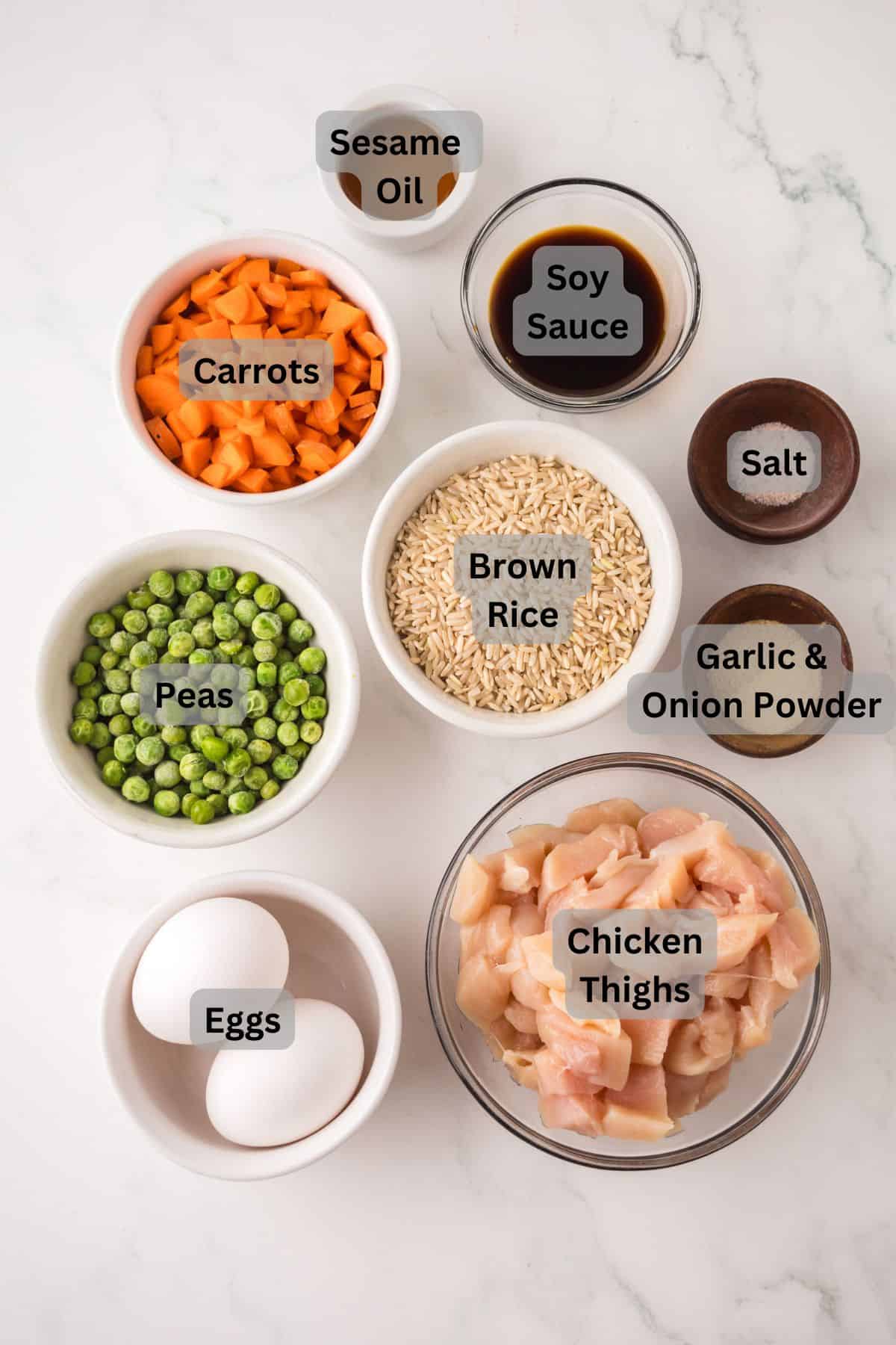 Top-down view of bowls containing labeled ingredients for making chicken fried rice: sesame oil, soy sauce, salt, carrots, garlic & onion powder, peas, brown rice, eggs, and diced chicken thighs on a white surface.