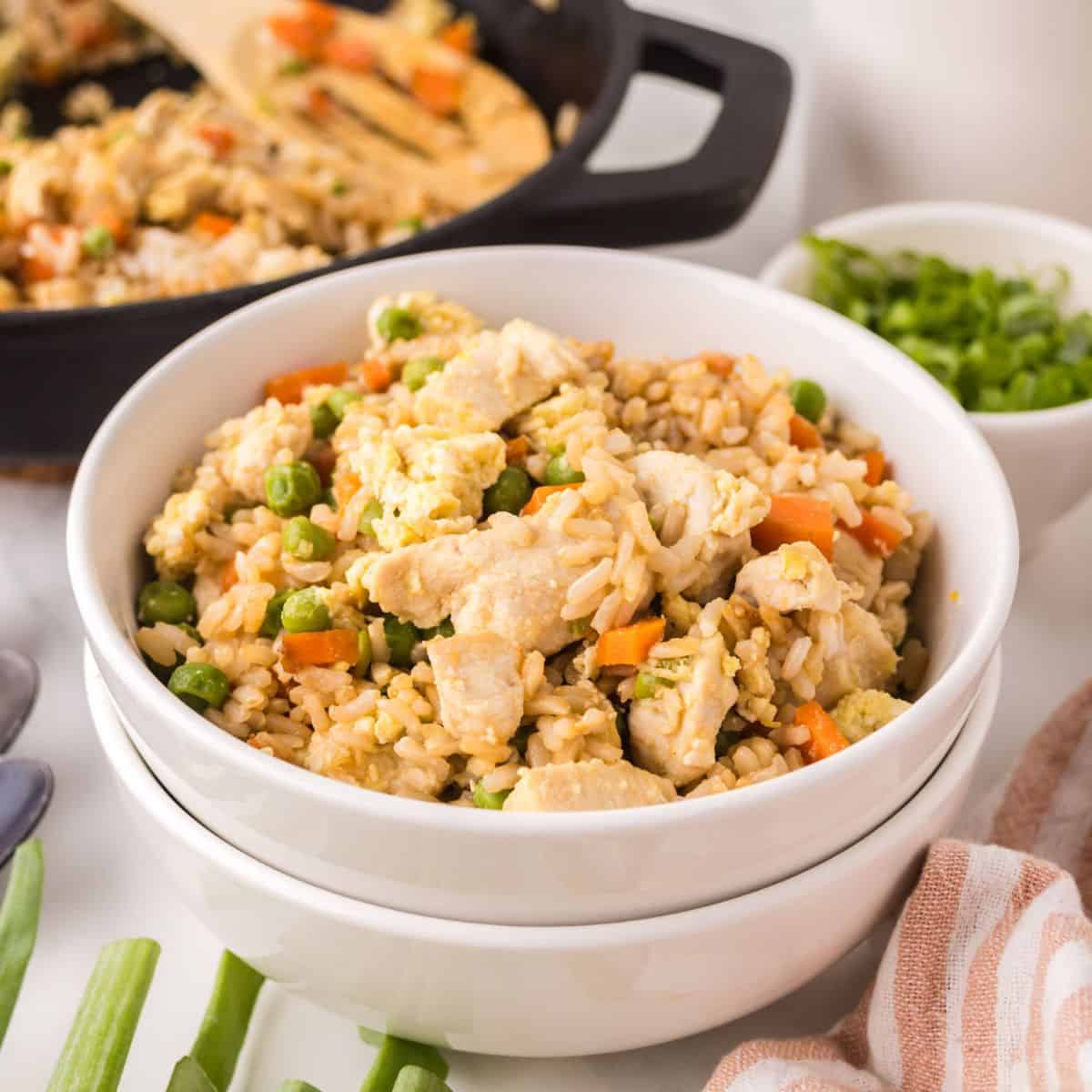 A bowl of chicken fried rice with peas, carrots, and scrambled eggs, served in a white bowl with a skillet and chopped green onions in the background—perfect for anyone making chicken fried rice at home.
