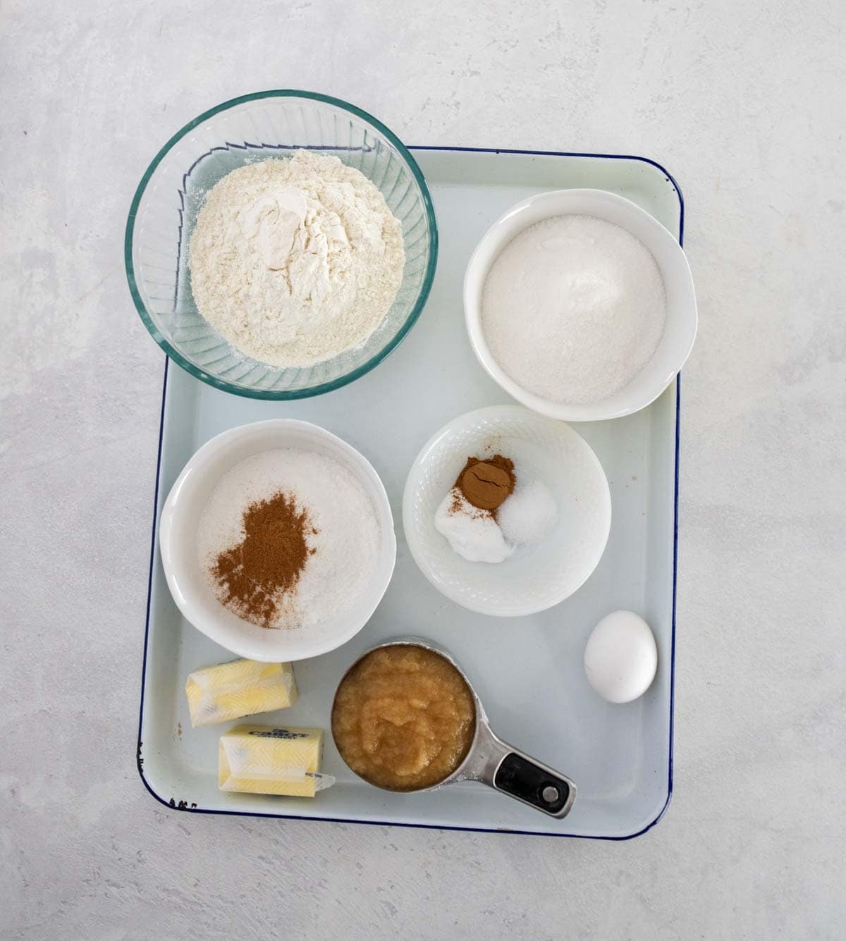 A tray with small bowls containing flour, sugar, cinnamon, baking soda, salt, and applesauce—perfect for baking an applesauce coffee cake—plus two butter sticks and a whole egg arranged neatly on a white surface.