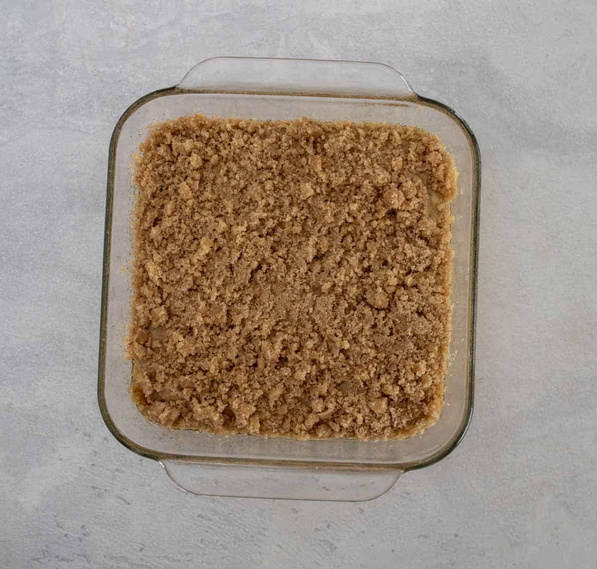 A square glass baking dish filled with a crumbly brown mixture, resembling unbaked streusel or crumb topping for an applesauce coffee cake, placed on a light gray surface.