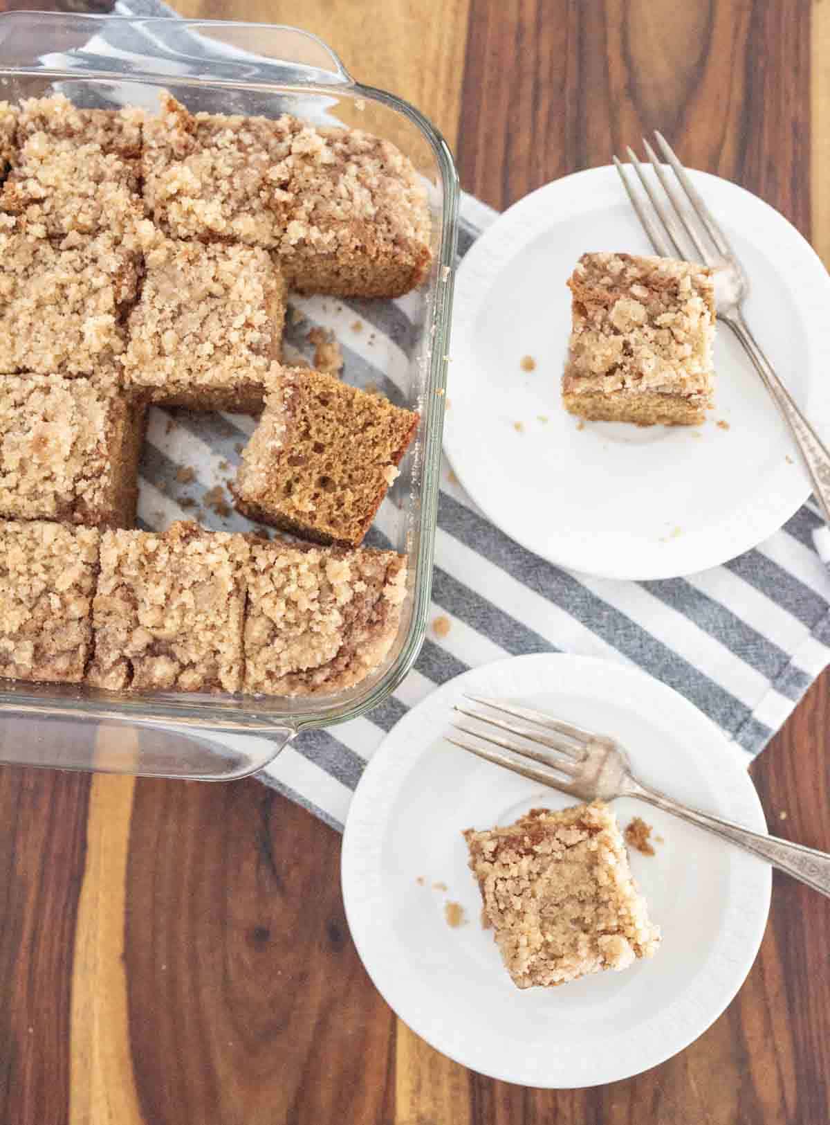 A glass baking dish with sliced applesauce coffee cake sits on a striped cloth. Two white plates each hold a piece of cake with a fork, displayed on a wooden table.