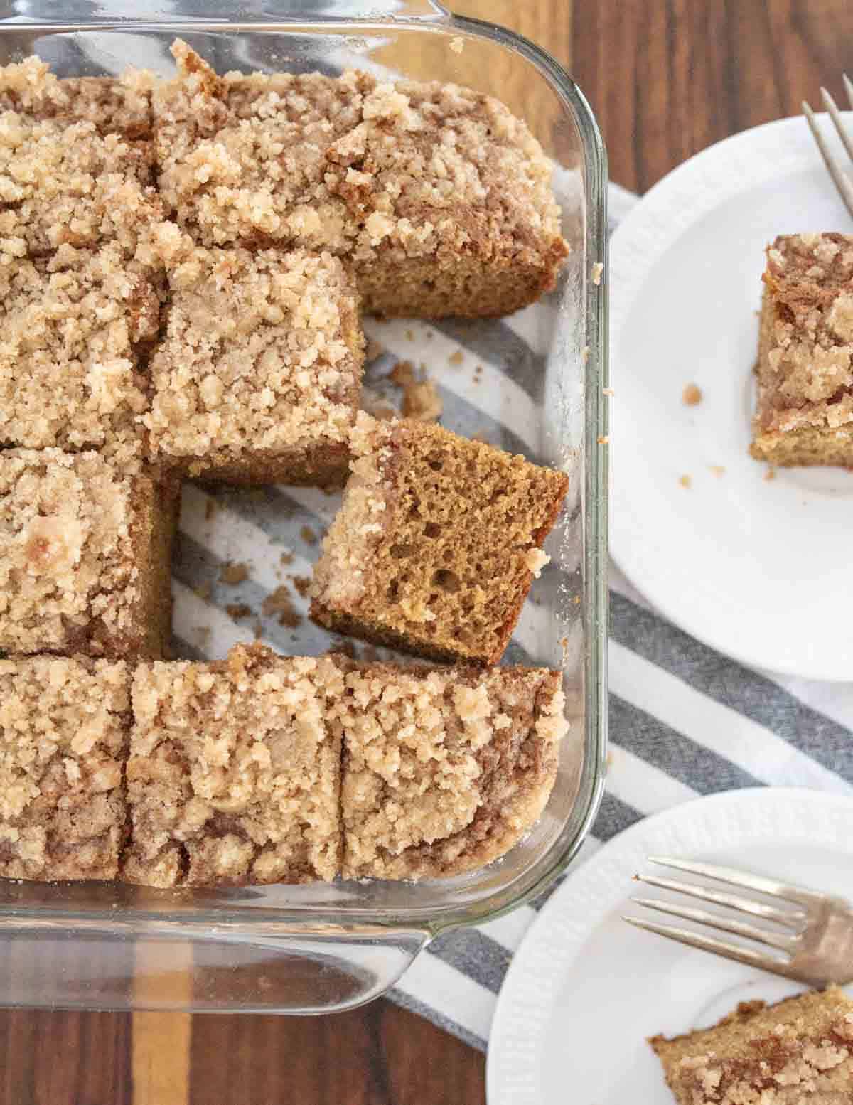 A glass baking dish filled with sliced pieces of crumb-topped applesauce coffee cake, with two pieces served on white plates beside it, each with a fork. The dish sits on a striped cloth on a wooden surface.