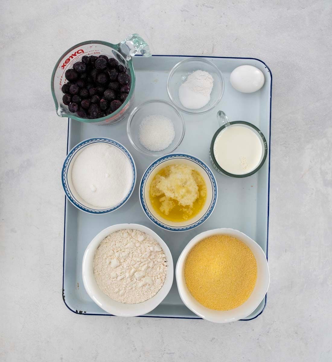 A tray with bowls and cups holding ingredients for blueberry cornbread—blueberries, flour, sugar, melted butter, cornmeal, baking powder, salt, milk, and one whole egg—arranged neatly on a light surface.