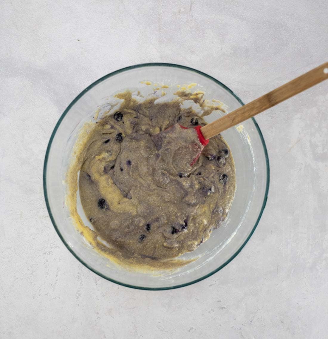 A glass bowl filled with blueberry cornbread muffin batter and a red spatula, sitting on a light gray surface.