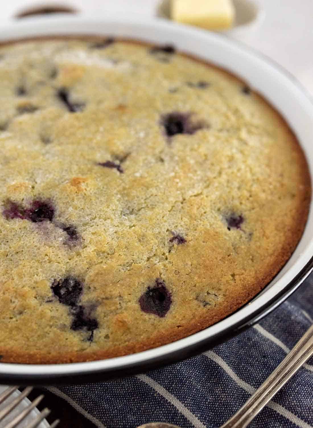 A close-up of freshly baked blueberry cornbread in a round white dish, its golden-brown crust dotted with juicy blueberries. A striped cloth and a blurred butter dish add cozy details to the background.