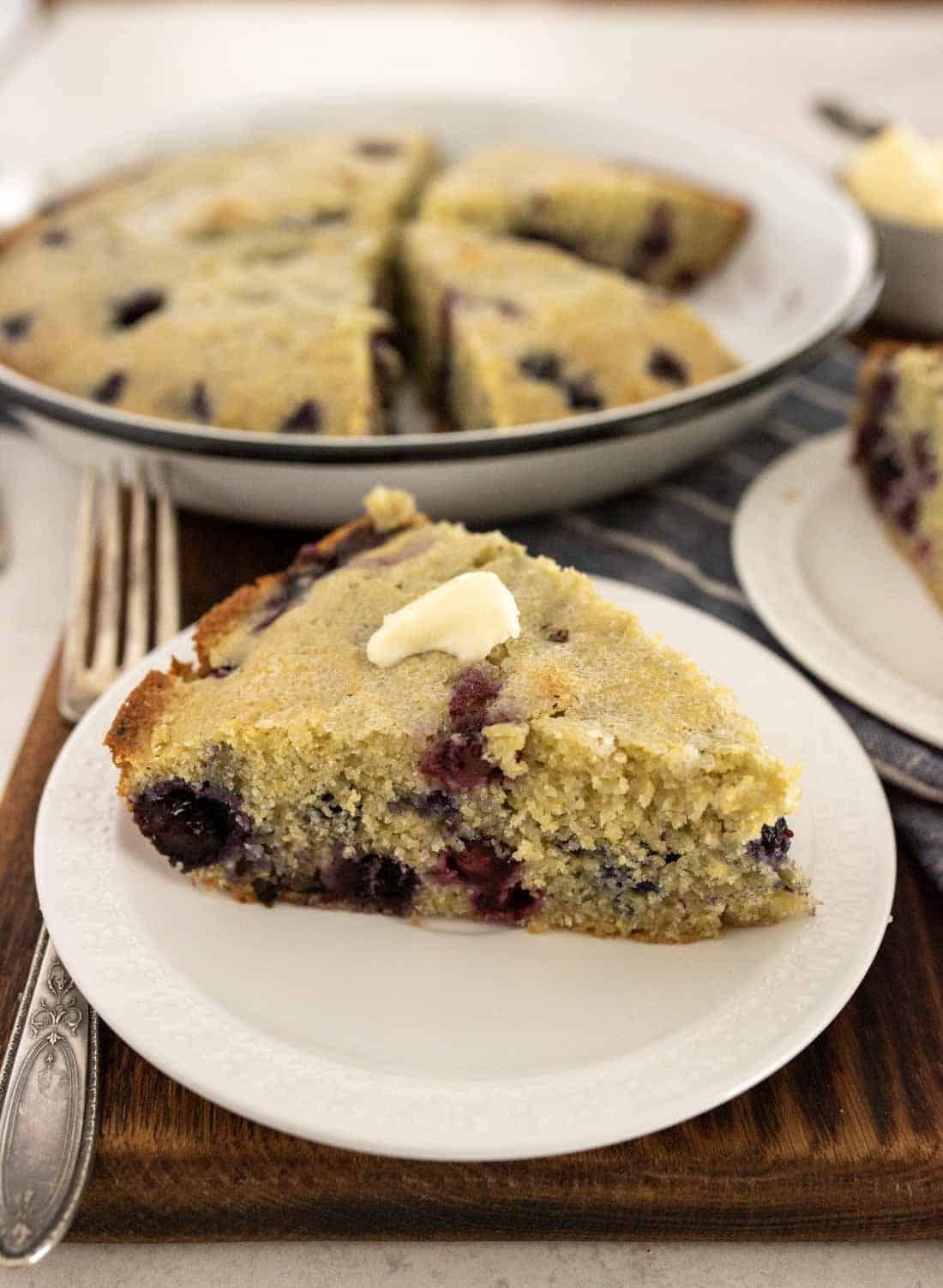 A slice of blueberry cornbread with a pat of butter on top sits on a white plate, the golden blueberry cornbread behind it cut into inviting wedges.