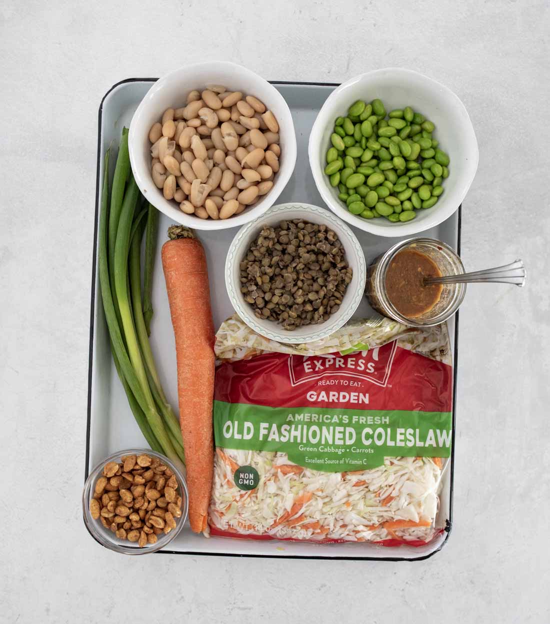 A tray holding a bag of coleslaw mix, a carrot, green onions, small bowls of white beans, edamame, lentils, crispy chickpeas, and a small bowl of salad dressing with a spoon.