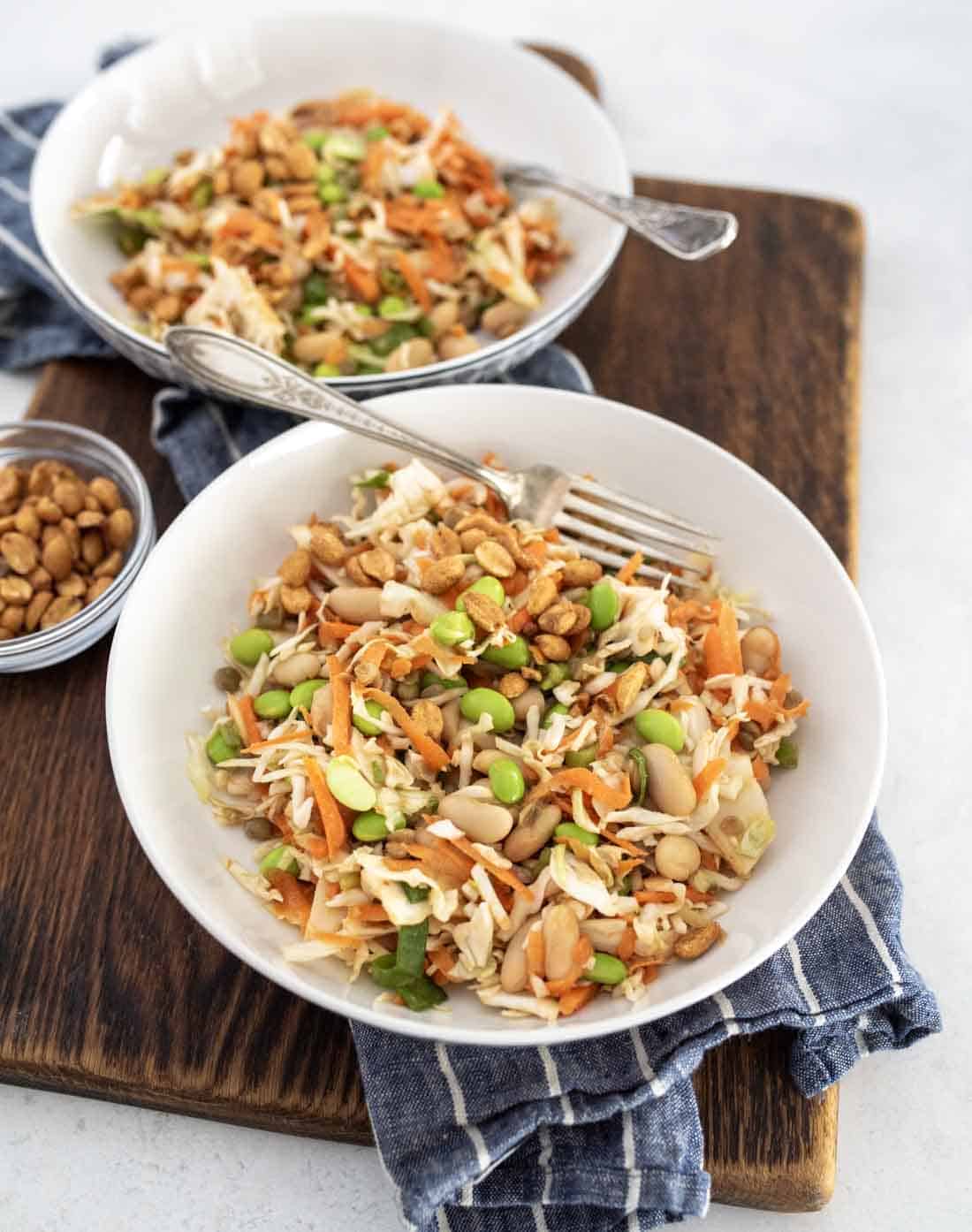 Two white bowls filled with colorful dense bean salad recipe containing shredded cabbage, carrots, edamame, white beans, and chopped peanuts, placed on a wooden board with a small bowl of peanuts and a blue checked cloth.