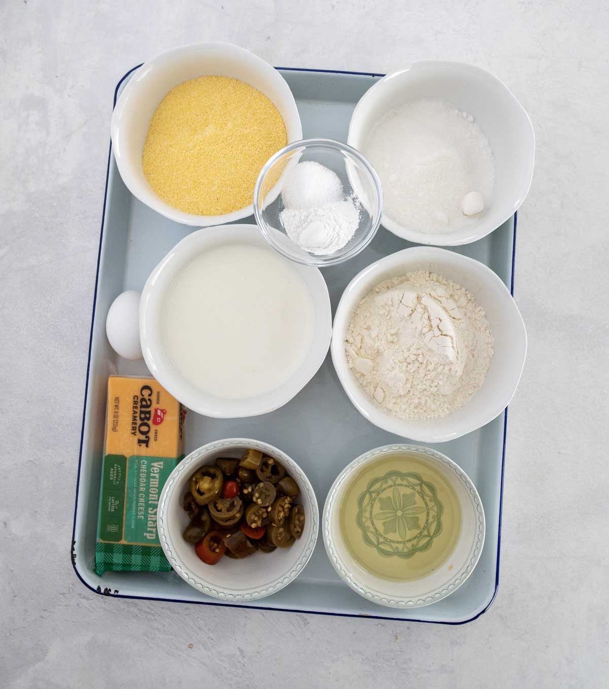 A tray with bowls containing cornmeal, sugar, flour, baking powder, milk, an egg, sliced jalapeños, a stick of butter, and a small bowl of oil, arranged on a light surface.