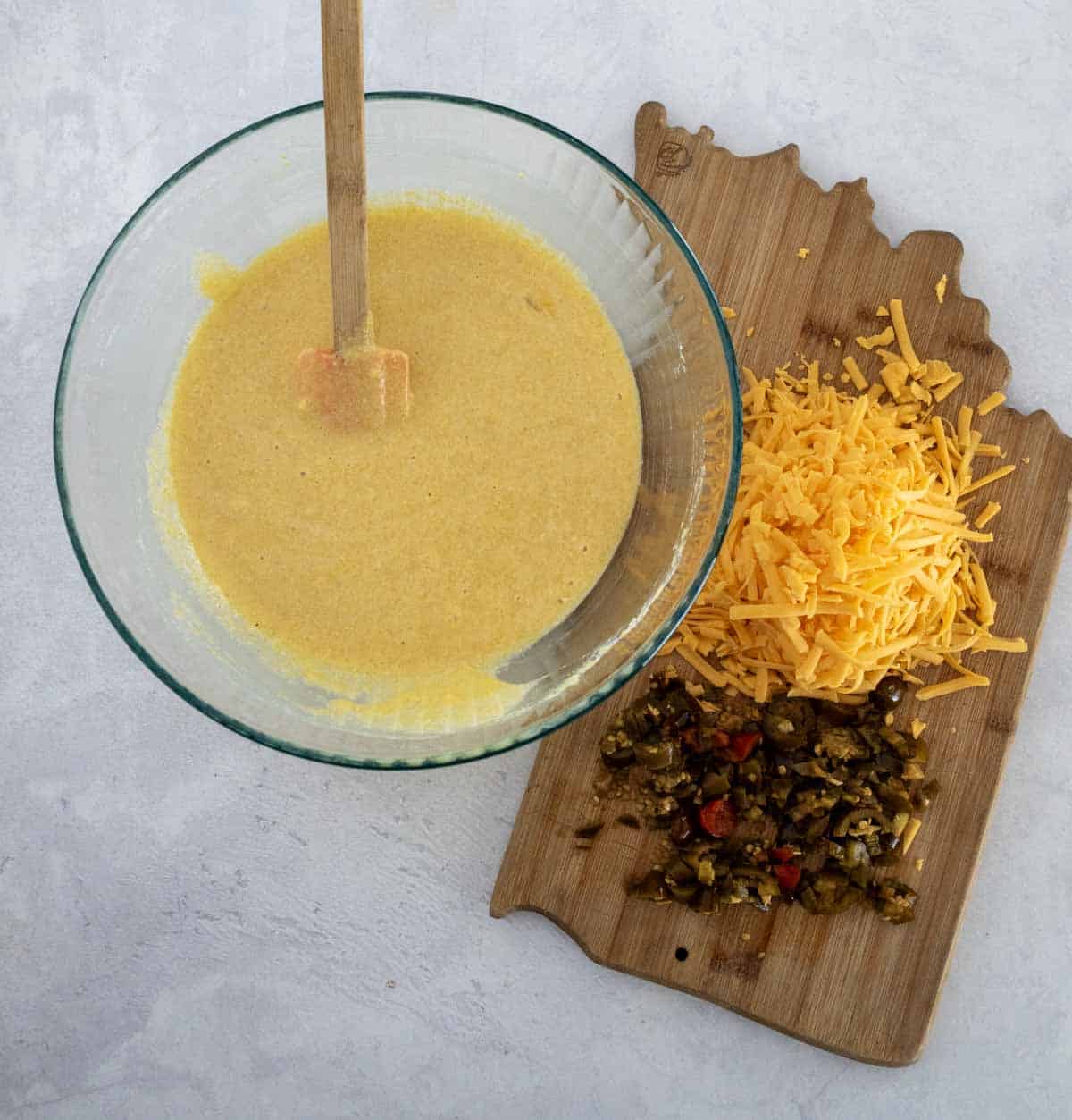 A glass bowl with cornmeal batter and a spatula next to a wooden board holding shredded cheddar cheese and chopped jalapeños on a light surface.