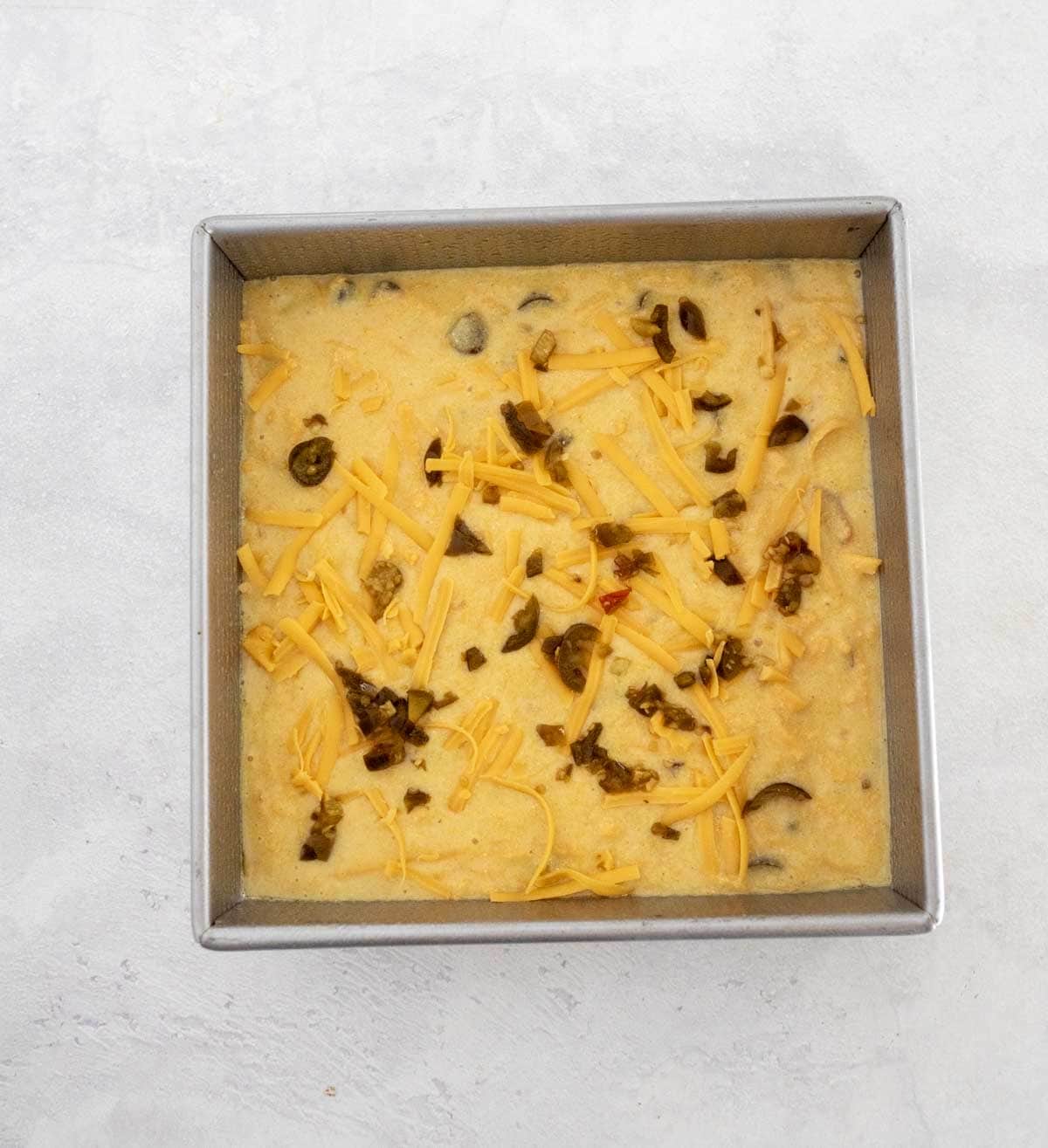 A square metal baking pan filled with unbaked cornbread batter, topped with shredded cheddar cheese and chopped jalapeños, on a light-colored surface.