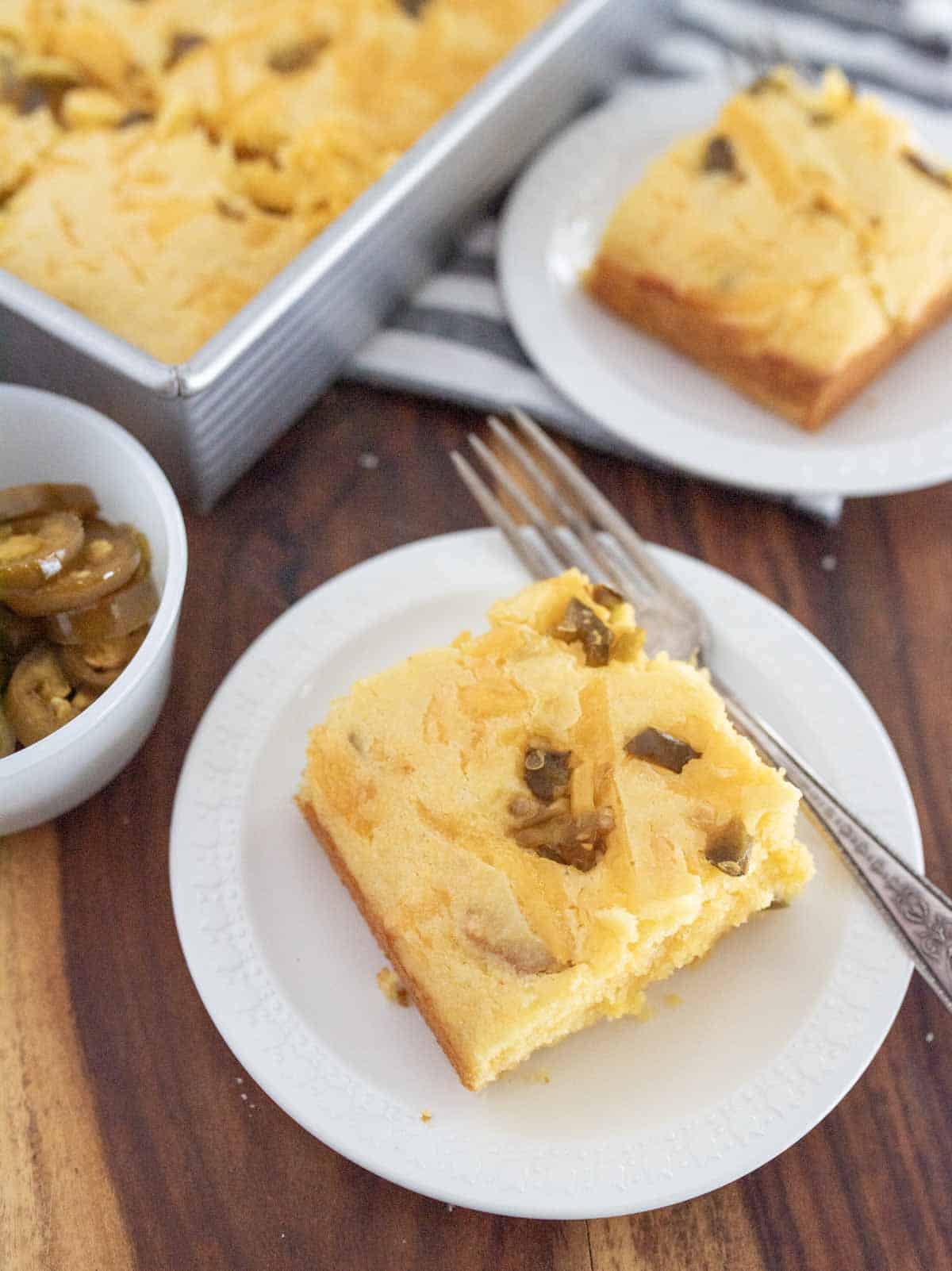 A slice of cornbread topped with jalapeños sits on a white plate with a fork. A pan of more cornbread and another plate with a slice are in the background, along with a small bowl of jalapeños.