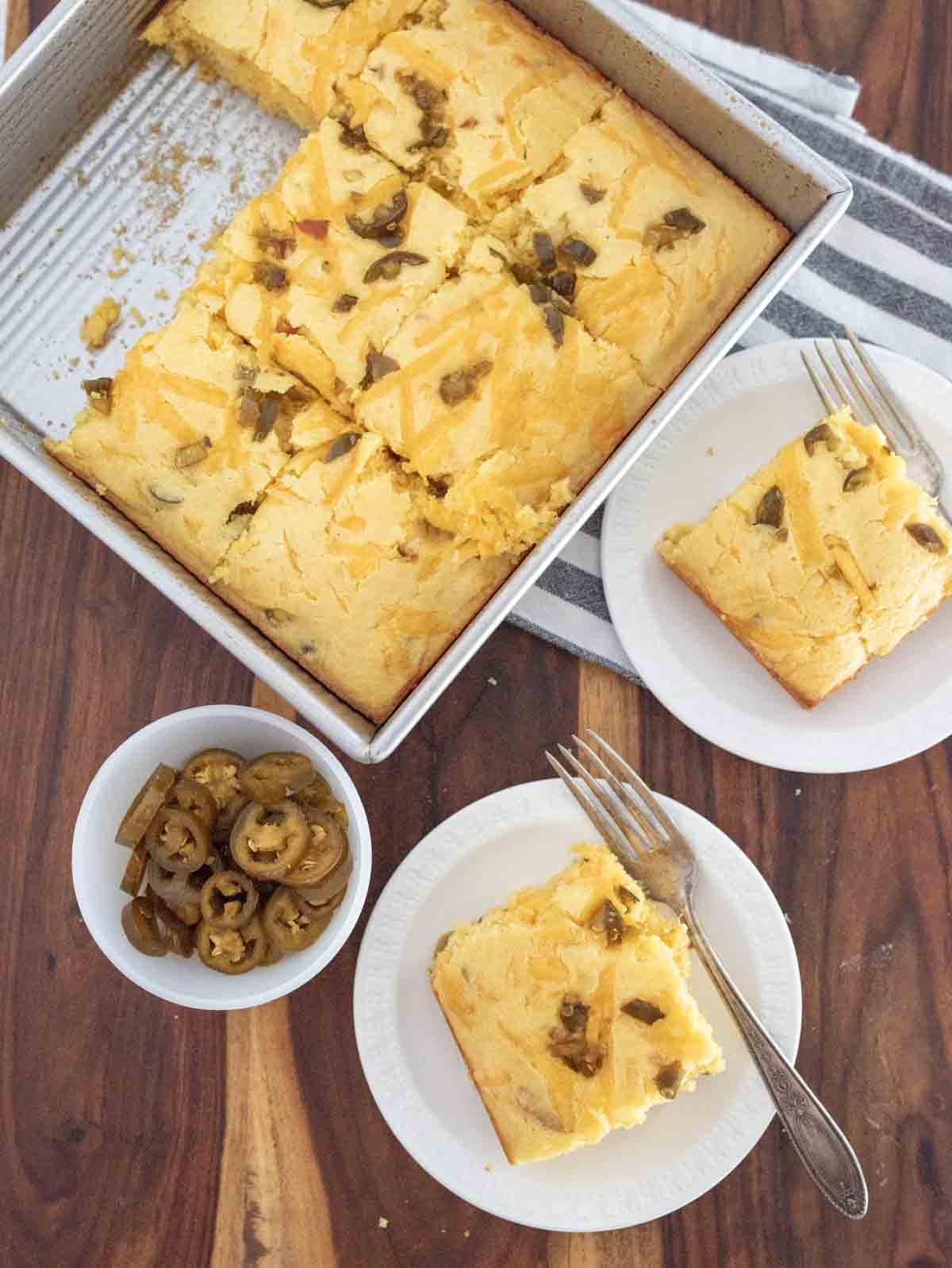 A square baking pan of cheddar and jalapeno cornbread sits on a wooden table. Two plates each hold a cheesy slice with a fork, and a small bowl contains pickled jalapeño slices.