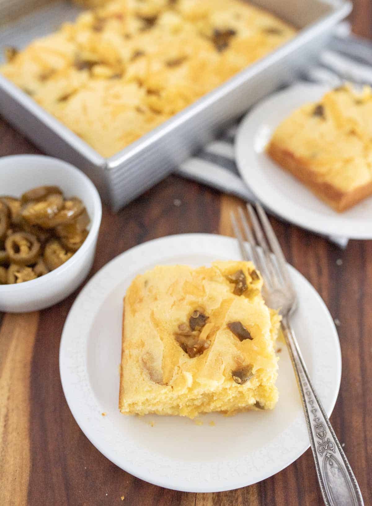 A slice of cornbread with jalapeños on a white plate with a fork, next to a bowl of sliced jalapeños and a pan of cornbread, all on a wooden table.