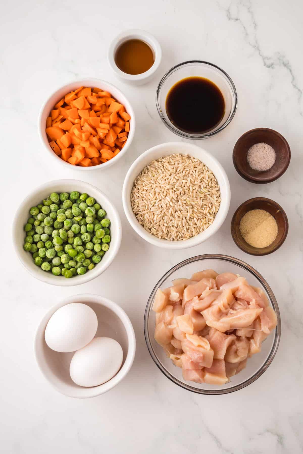 A top-down view of ingredients in bowls on a white surface, perfect for making chicken fried rice: raw diced chicken, eggs, peas, carrots, rice, soy sauce, oil, salt, and garlic powder.