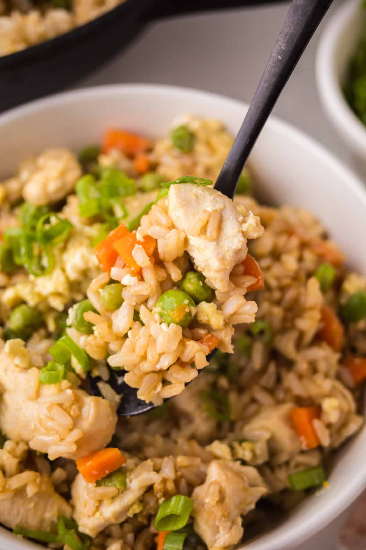 A close-up of a bowl of chicken fried rice with peas, carrots, scrambled eggs, and green onions, showing a spoon lifting a portion—perfect for anyone interested in making chicken fried rice at home.