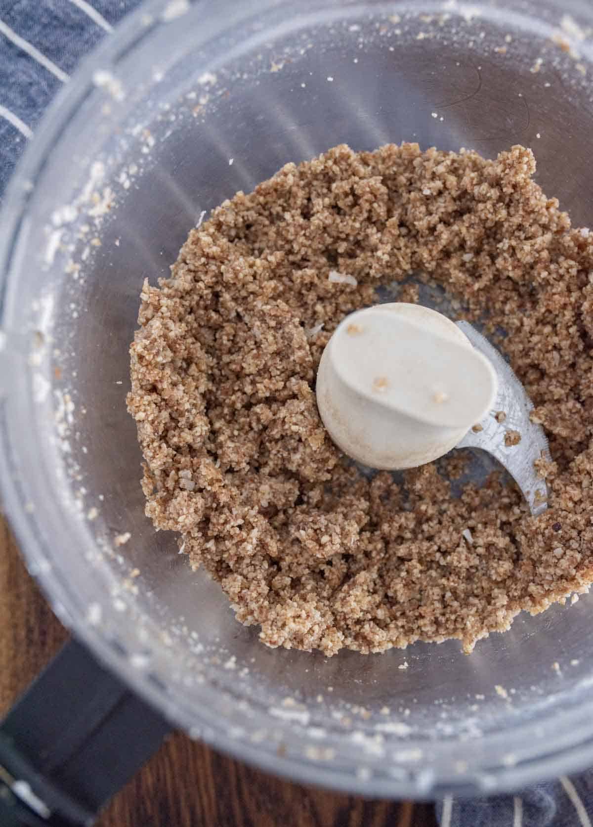 A top-down view of a food processor containing a coarse, crumbly mixture of blended nuts and ingredients, sitting on a wooden surface next to a striped cloth.
