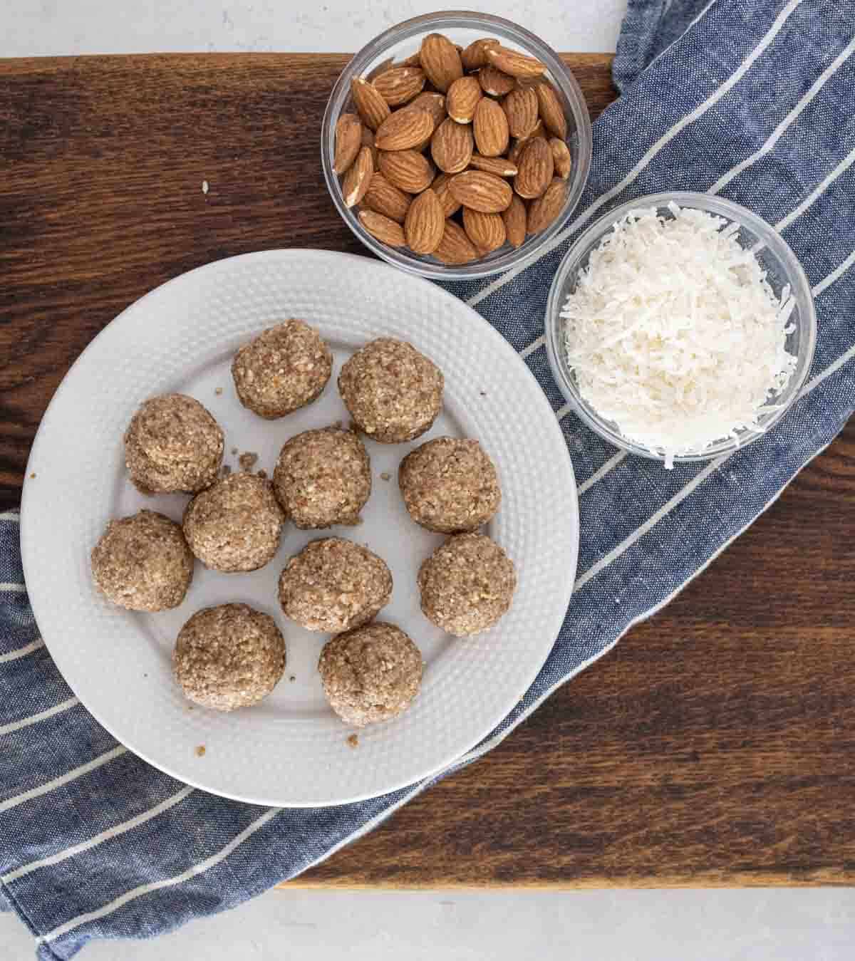 A white plate with ten almond energy balls sits on a wooden surface near two bowls—one with whole almonds and one with shredded coconut—on top of a blue-and-white striped cloth.