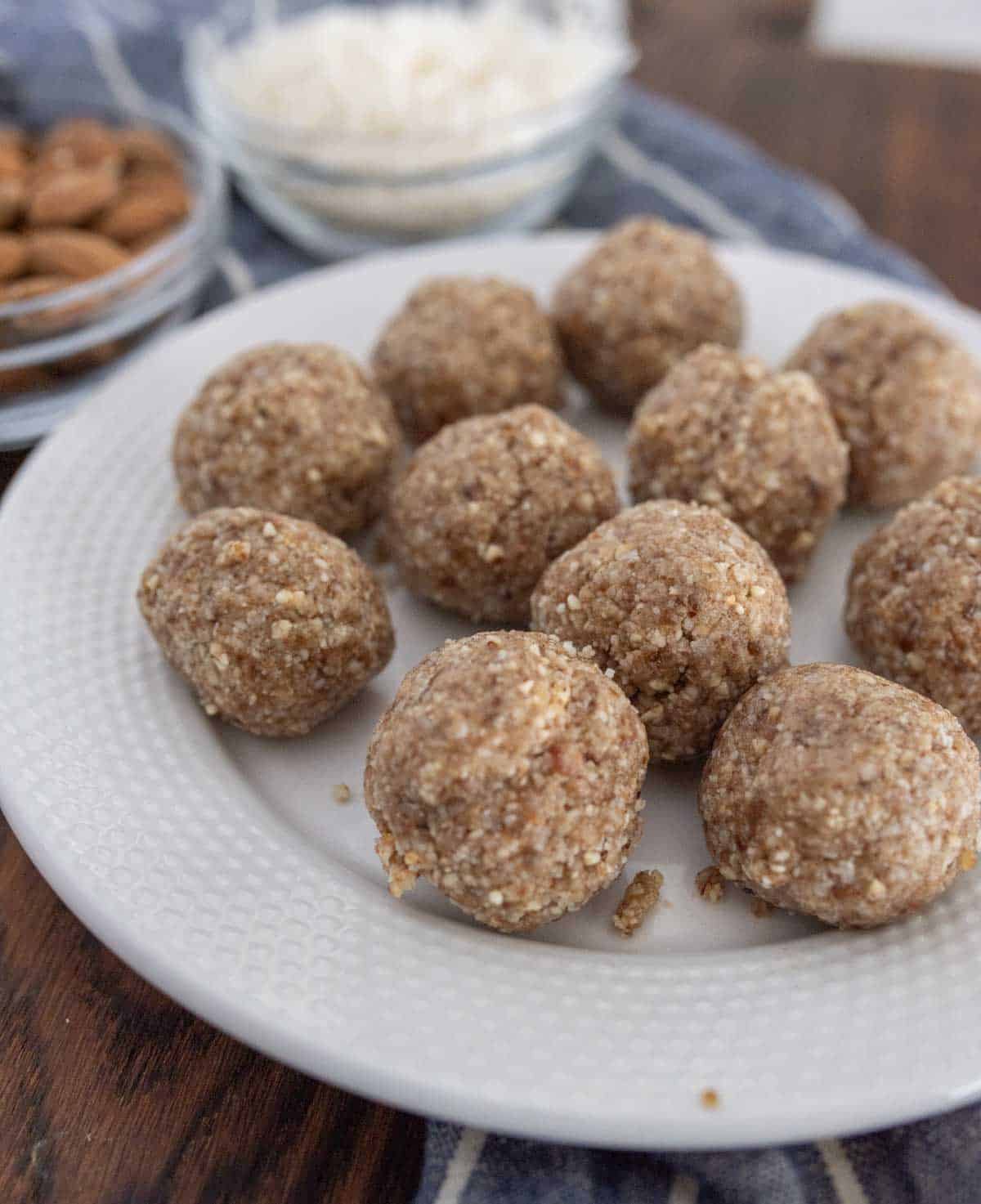A white plate with several round, brown coconut date nut balls on it. In the background are small glass bowls containing shredded coconut and whole almonds, sitting on a wooden surface.