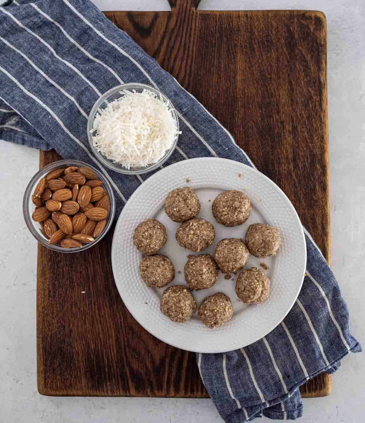 A white plate with ten round energy bites, a bowl of almonds, and a bowl of shredded coconut sit on a wooden cutting board with a blue-striped kitchen towel underneath.