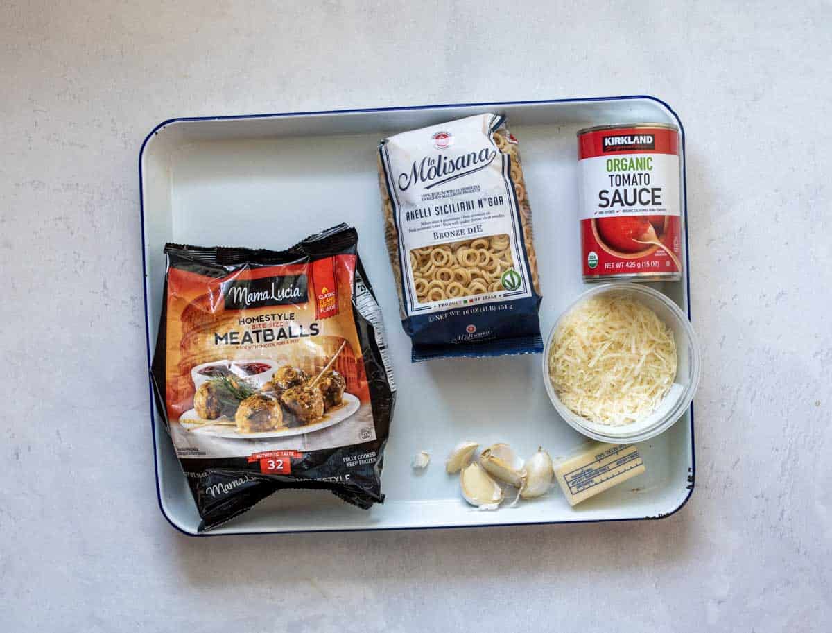 A tray with a bag of frozen meatballs, a pack of pasta, a box of tomato sauce, a container of shredded cheese, and several garlic cloves arranged on a white surface.