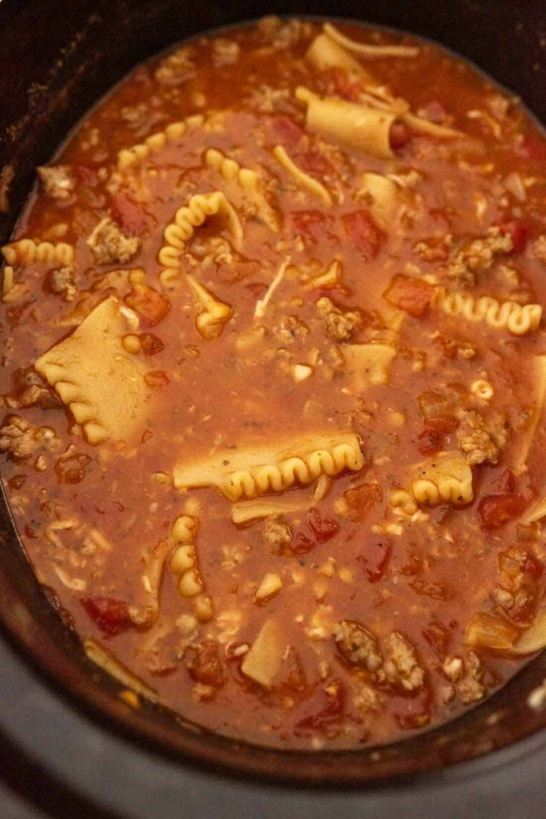 A close-up of lasagna soup in slow cooker, showing broken lasagna noodles, ground meat, diced tomatoes, and a rich tomato-based broth.