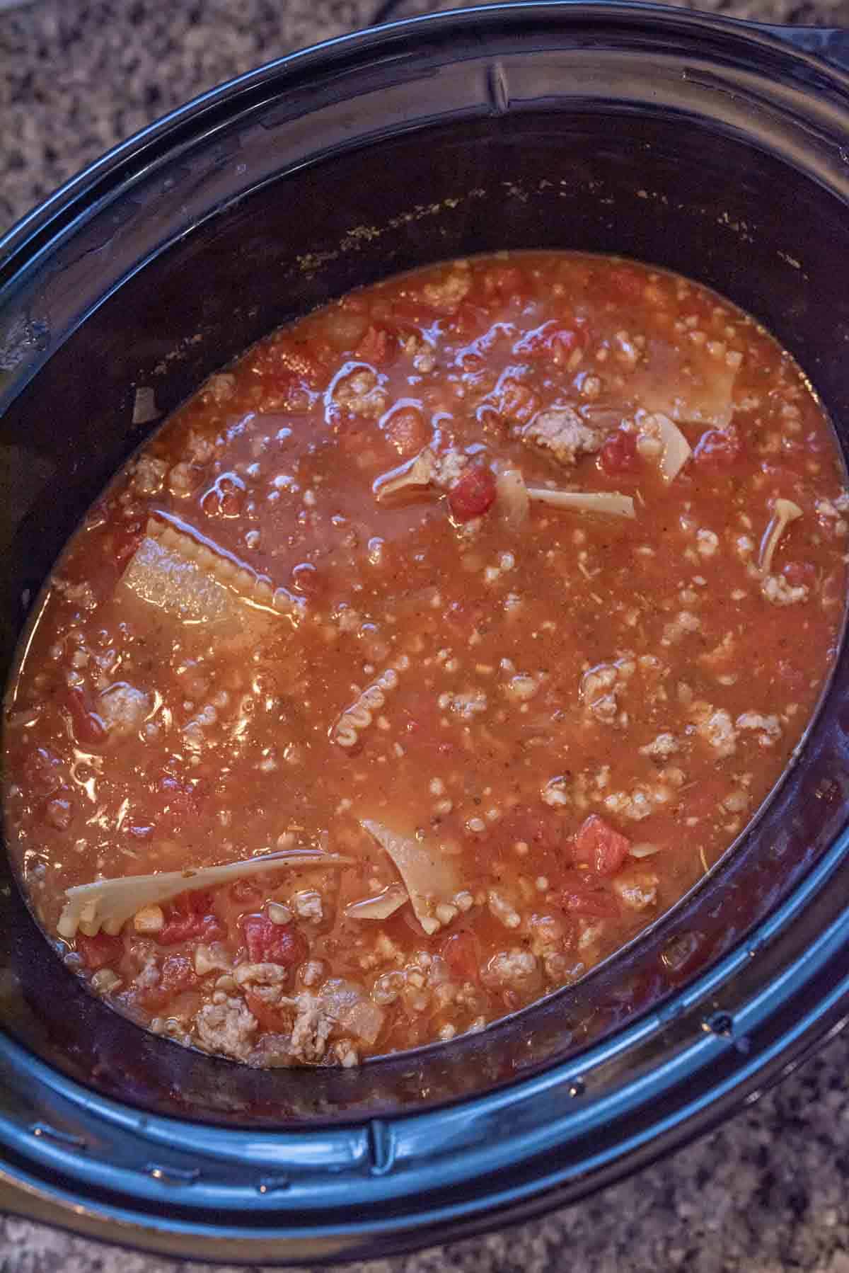 A slow cooker filled with lasagna soup in slow cooker, featuring ground meat, tomato sauce, diced tomatoes, and broken lasagna noodles on a granite countertop.
